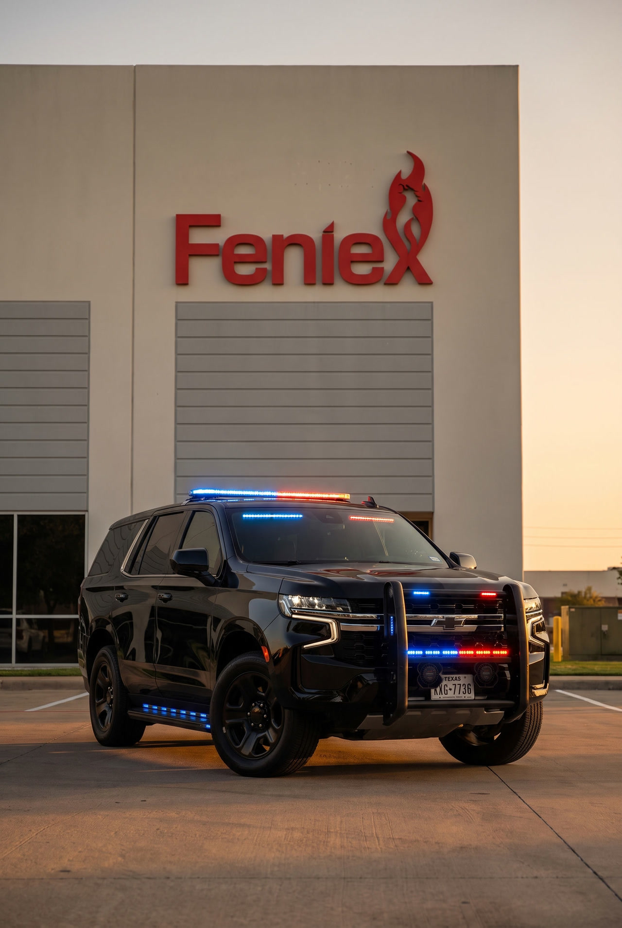 A black Chevrolet Tahoe fitted with Feniex lighting, lightbar, grille and running-board lights lit red and blue, parked outside the Feniex Industries building in Austin, Texas at sunset.